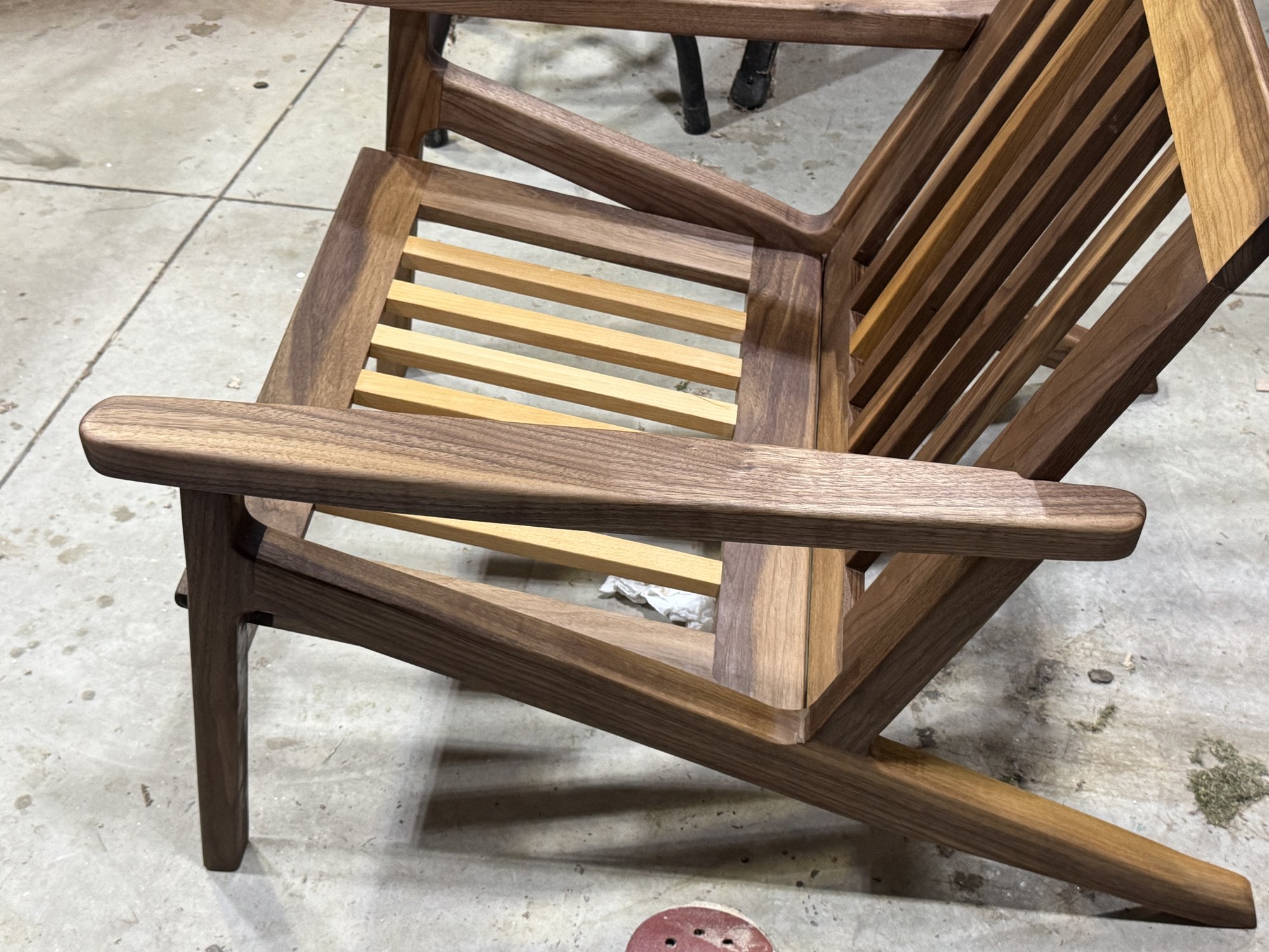 Workshop three-quarter view of the unfinished-looking walnut chair frame, highlighting a wide arm, seat slats, and slatted back.