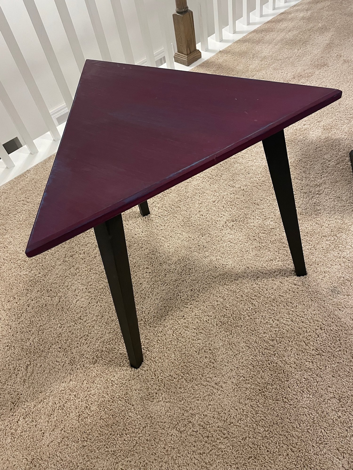 Three-quarter view of the triangular poplar table standing on the carpet, plum top and black tapered legs.
