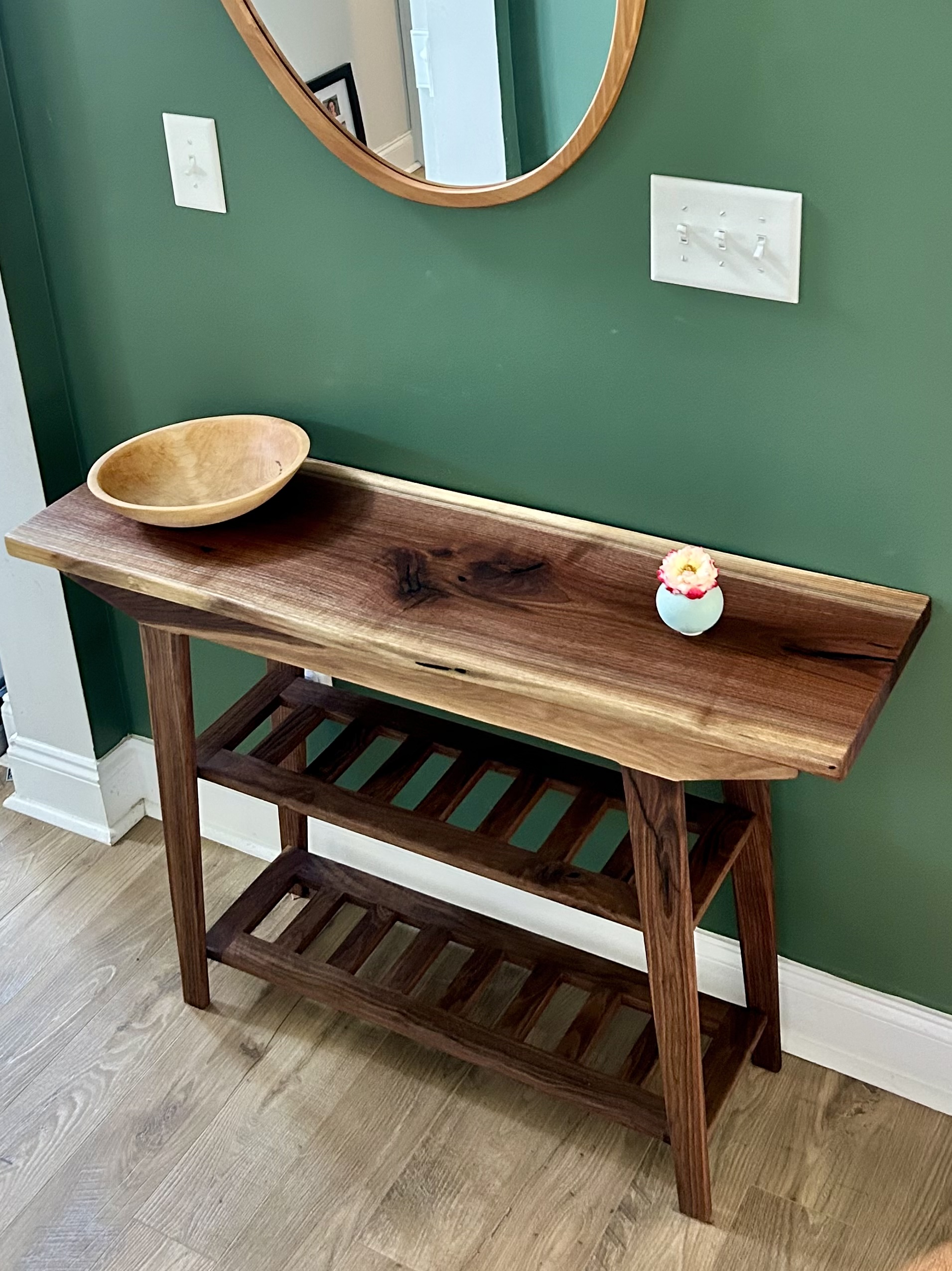 Straight-on view of the finished walnut console and shoe rack against a green wall, with the wooden bowl and small vase on the live-edge top.